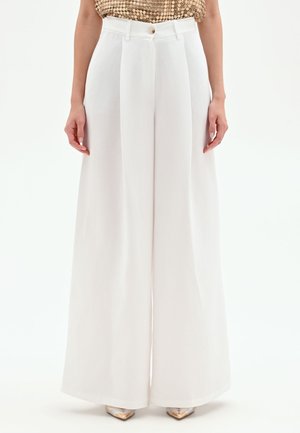Pantalon blanc taille haute à jambes larges avec plis devant, taille avec bouton, porté avec un top à sequins métalliques et des talons pointus.