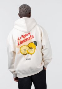 Uomo con capelli grigi ricci che indossa un hoodie color crema con una grafica di limone e il testo rosso "La Migliore Limonata" sulla schiena, con le mani nelle tasche.