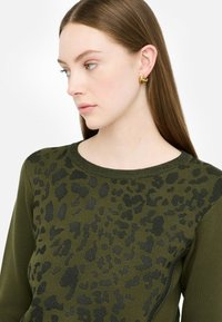 Olive roheline sveter musta leopardimustriga, ümmargune kaelus, ribakujundusega servad ja sujuv tekstuur, millele lisavad sära kuldsed kõrvarõngad.