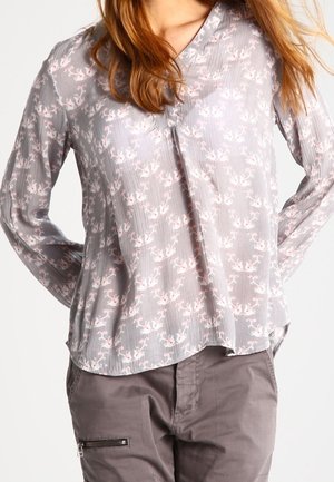 Femme portant un chemisier à manches longues gris clair à motifs floraux, rentré dans un pantalon gris avec une poche zippée à l'avant, debout les mains derrière le dos.