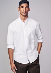 Camicia bianca a maniche lunghe con maniche arrotolate e taschino sul petto, realizzata in un tessuto liscio, indossata con pantaloni marroni scuri.