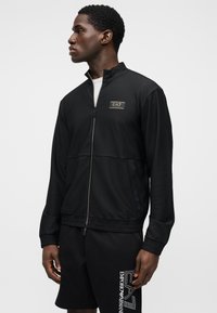 Melna rāvējslēdzēja jaka ar teksturētu audumu, augstu apkakli un logotipa uzšuvumu. Raksturīgas elastīgas aproces un pieguloša dizaina siluets.