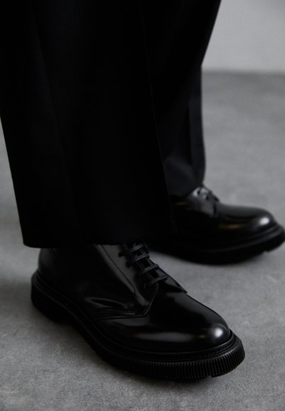 Zwarte lederen schoenen met een glanzende afwerking, voorzien van een ronde neus en veters. De zool is gestructureerd en licht verhoogd, wat perfect past bij op maat gemaakte broeken.