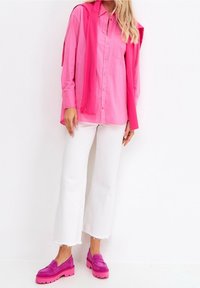 Kvinna som bär en ljusrosa skjorta med knappar och en matchande tröja över axlarna, vita capribyxor och rosa platåskor.