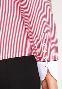 Manga de camisa a rayas rojas y blancas con un puño blanco liso que presenta tres botones circulares y un ribete en contraste de color marino.