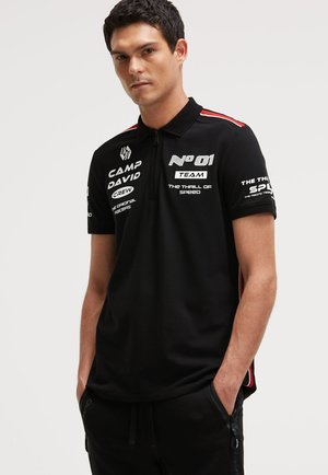 Young man wearing a black short-sleeve polo with white racing-themed text and red stripe accents, hands in pockets, neutral background.
