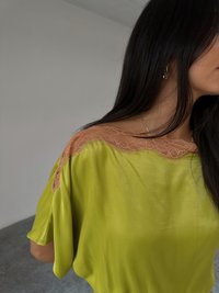 En kvinne med langt mørkt hår har på seg en lys grønn topp med en gjennomsiktig blonde halsringning, som viser et minimalistisk interiør med et glatt gulv.