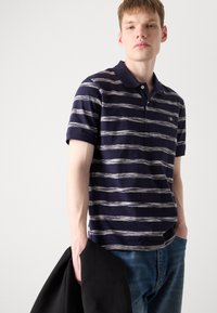 Marine stripete polo-skjorte med hvite horisontale linjer, kortermet og klassisk krage. Modellen holder en svart jakke i den ene hånden.