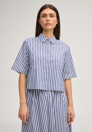 Frau mit glatten braunen Haaren, die ein blau-weiß gestreiftes kurzärmeliges Hemd und einen passenden Rock trägt, vor einem einfarbigen Hintergrund steht.