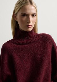 Jersey de cuello alto burdeos hecho de material de punto grueso con un cuello acanalado, que presenta un patrón texturizado y un corte holgado.