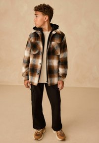 Checkered flannel jacket in brown, black, and white; black hoodie underneath, cream t-shirt, black cargo pants, and light brown sneakers.