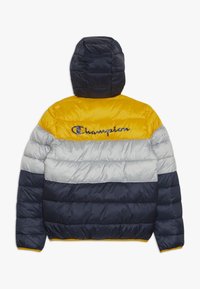 Pufferjackan har en färgblockdesign i marinblått, grått och gult, med huva och broderad "Champion"-logotyp på ryggen.