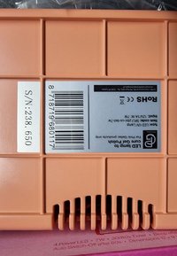 Vierkant, lichtoranje plastic omhulsel met ventilatiesleuven. Een wit etiket toont elektrische specificaties en een streepjescode.