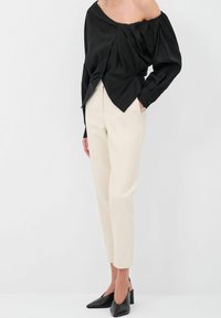 Blusa nera off-the-shoulder con frontale drappeggiato e dettagli con bottoni, abbinata a pantaloni beige sartoriali e mule nere con tacco.