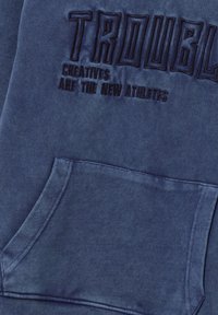 Felpa blu navy con ricamo della scritta "TROUBLE" e la frase "I CREATIVI SONO I NUOVI ATLETI", con tasca frontale e tessuto testurizzato.