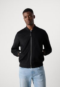 Black bomber jacket in soft fabric with a zip front, ribbed cuffs, and two side pockets. Paired with light blue denim jeans.