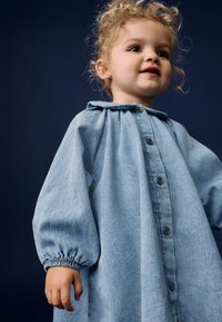 Hellblaues Denimkleid mit langen, gepufften Ärmeln, durchgehender Knopfleiste und Kragen. Der Stoff wirkt weich mit einer leicht abgetragenen Textur.