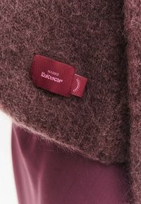 Ciemnobordowy dzianinowy sweter o miękkiej, puszystej fakturze. Z czerwonym nadrukiem „NAOKO Knitwear” w różowej czcionce. Widoczny owinięty materiał.