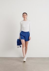 Camisa branca de mangas compridas, calções azuis e ténis brancos. Segurando uma mochila azul e cinza, destacando-se num fundo claro.