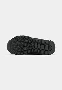 Semelle de chaussure en caoutchouc noir avec des sculptures texturées carrées et rectangulaires pour une meilleure adhérence, vue depuis le dessous sur un fond clair.