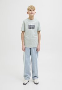 Ljusgrön t-shirt med kort ärm som har en rektangulär svart och vit grafik på framsidan, i kombination med ljusblå vida jeans och grå sneakers.