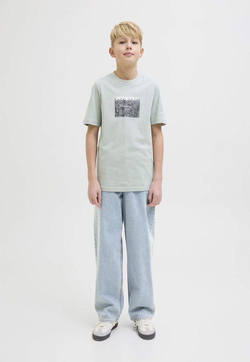 Ljusgrön t-shirt med kort ärm som har en rektangulär svart och vit grafik på framsidan, i kombination med ljusblå vida jeans och grå sneakers.