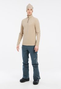 Pullover in pile beige con collo alto e design a mezza zip, abbinato a pantaloni waterproof blu scuro con tasche con zip. Stivali neri.