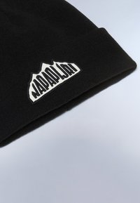 Czarna czapka beanie wykonana z miękkiego materiału dzianinowego, z białą łatką z logo, na której znajdują się grafiki gór oraz tekst, umieszczona na złożonym brzegu.