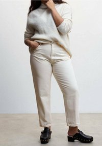 Personne portant un pull en tricot crème, un pantalon écru taille haute et des sabots noirs ornés de clous dorés, debout devant un fond neutre.