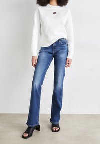White sweatshirt with small graphic, paired with blue flared jeans and black heeled sandals. Smooth texture, casual design, and contrasting colors.