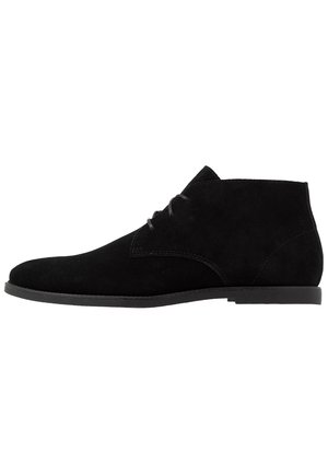 Bottine noire en daim à lacets sur le devant, à bout arrondi et petit talon en caoutchouc, représentée de profil sur fond blanc.