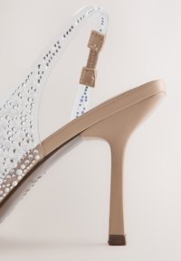 Escarpin beige à talon haut avec une bride transparente décorée de petits clous ronds argentés et blancs et deux boucles de fixation beiges.