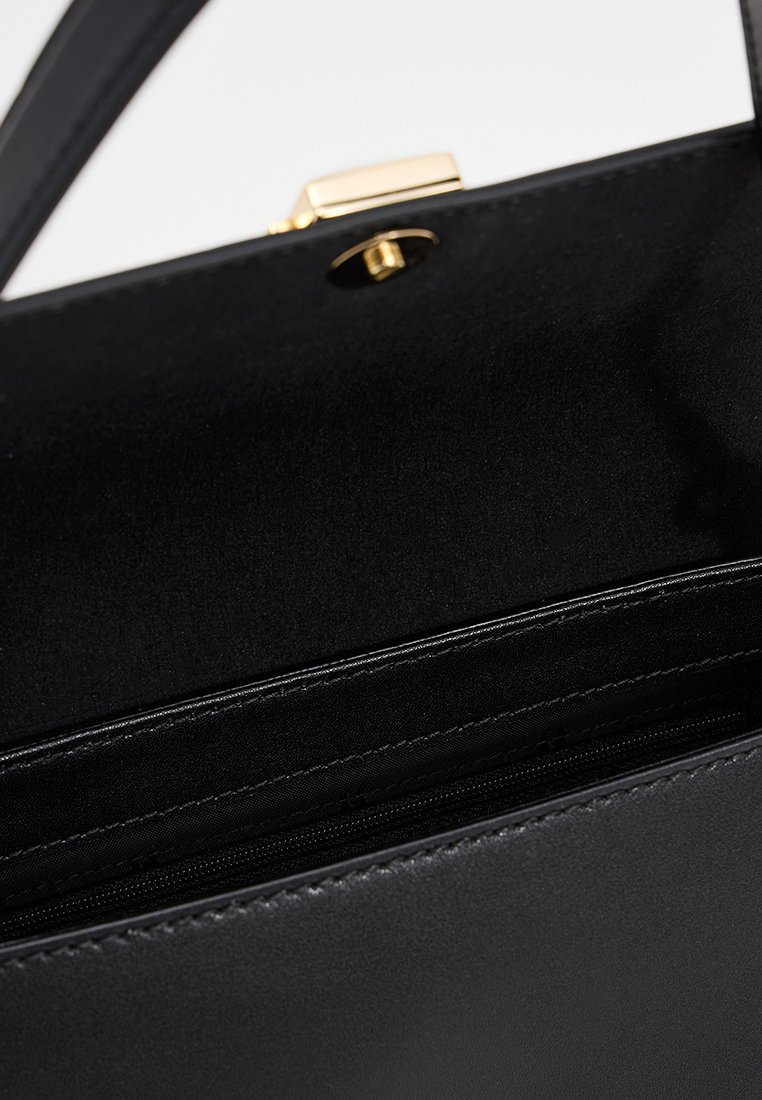 Intérieur d'un sac à main en cuir noir montrant une poche zippée et un fermoir en ton doré sur le rabat ouvert.