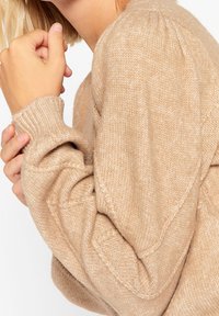 Beige stickad tröja med ribbade ärmslut, mjuk textur och avslappnad passform. Har diskreta detaljer på ärmarna och en mysig utseende.