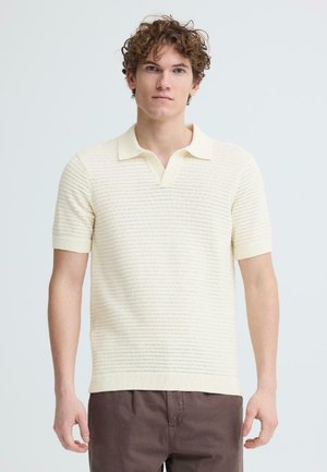 Junger Mann mit lockigem Haar, der ein cremefarbenes, kurzärmeliges Strick-Poloshirt und braune Hosen trägt, vor einem schlichten hellen Hintergrund steht.