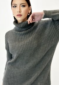 Jersey de cuello alto gris con canalé, hecho de un material suave. Presenta un cuello alto y un corte holgado con mangas largas. Textura sutil en la tela.