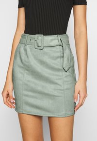 Grön konstmocka mini kjol med bälte i midjan, en jämn textur och ren sömnad längs fållen.