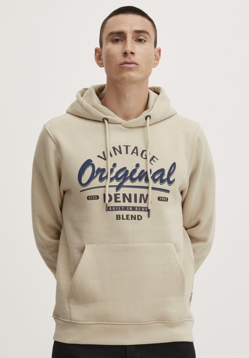 Beige hoodie with a kangaroo pocket, featuring navy blue printed text "Vintage Original Denim" and drawstrings on the hood. Soft fabric.