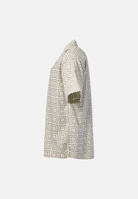 Chemise beige à manches courtes avec des motifs carrés et des points noirs, vue de côté sur un fond uni.