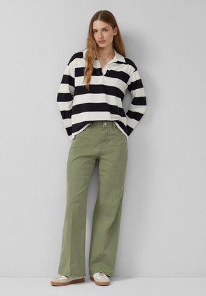 Frau trägt ein langärmliges schwarz-weiß gestreiftes Oberteil, olivgrüne Hosen mit weitem Bein und weiße Sneakers, steht vor einer schlichten grauen Wand.