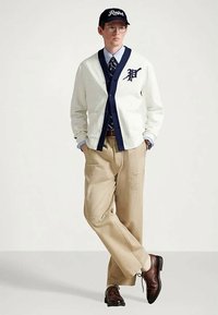 Άνδρας με γυαλιά και καπέλο, φορώντας λευκό κασκόλ με ναυτική επένδυση, ριγέ πουκάμισο, γραβάτα, μπεζ παντελόνι και καφέ κοστούμι, στέκεται με τα χέρια στις τσέπες.