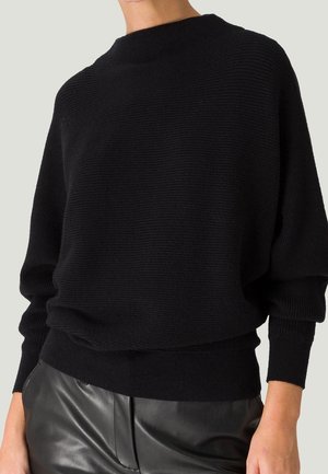 Pull côtelé noir avec manches chauve-souris associé à un pantalon en cuir noir, montré sur une personne du cou aux hanches.