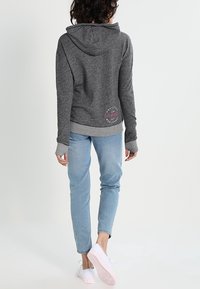 Grå huvtröja med en ljusgrå insidan, med en logotypdetalj på nedre delen av ryggen, i kombination med ljusblå jeans och vita skor.