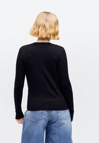 Άτομο με κοντά ξανθά μαλλιά που φοράει μαύρο μπλουζάκι με μακριά μανίκια και ανοιχτόχρωμο τζιν, φαινόταν από πίσω σε λευκό φόντο.