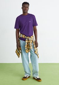 T-shirt roxa de algodão com mangas curtas, vestida sobre uma camisa de flanela amarela e em padrão xadrez atada à cintura. Jeans de ganga azul claro e ténis castanhos.