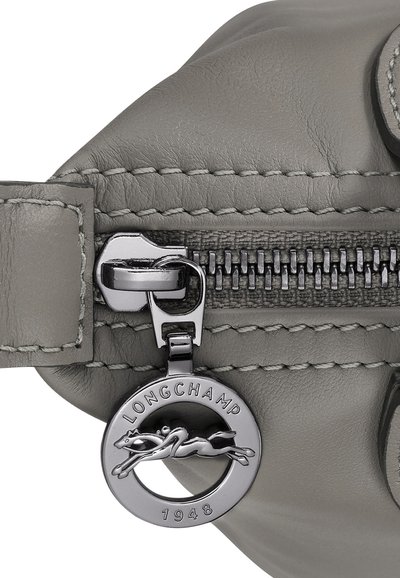 Detail einer grauen Ledertasche mit einem silbernen Reißverschluss, gestickten Akzenten und einem runden silbernen Anhänger mit einem laufenden Pferd und der Gravur „LONGCHAMP 1948“.