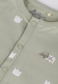 Body in cotone verde chiaro con motivi a corona bianchi, bottoni frontali e logo ricamato in marrone. Tessuto testurizzato con finitura liscia.