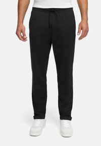 Svart joggebukse laget av myk stoff, med elastisk midjebånd med snøring, rett snitt og sidelommer. Brukt med hvite joggesko.