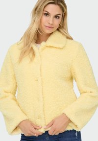 Yellow fluffy jacket with a textured surface, button closure, wide sleeves, and a collared neck. Worn over a light top and jeans.
