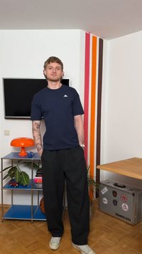 Mladić s tetovažama stoji u sobi s televizorom montiranim na zid, šarenim prugastim zidom, narančastom lampom, biljkama i koferom s natpisom "Tropicana".